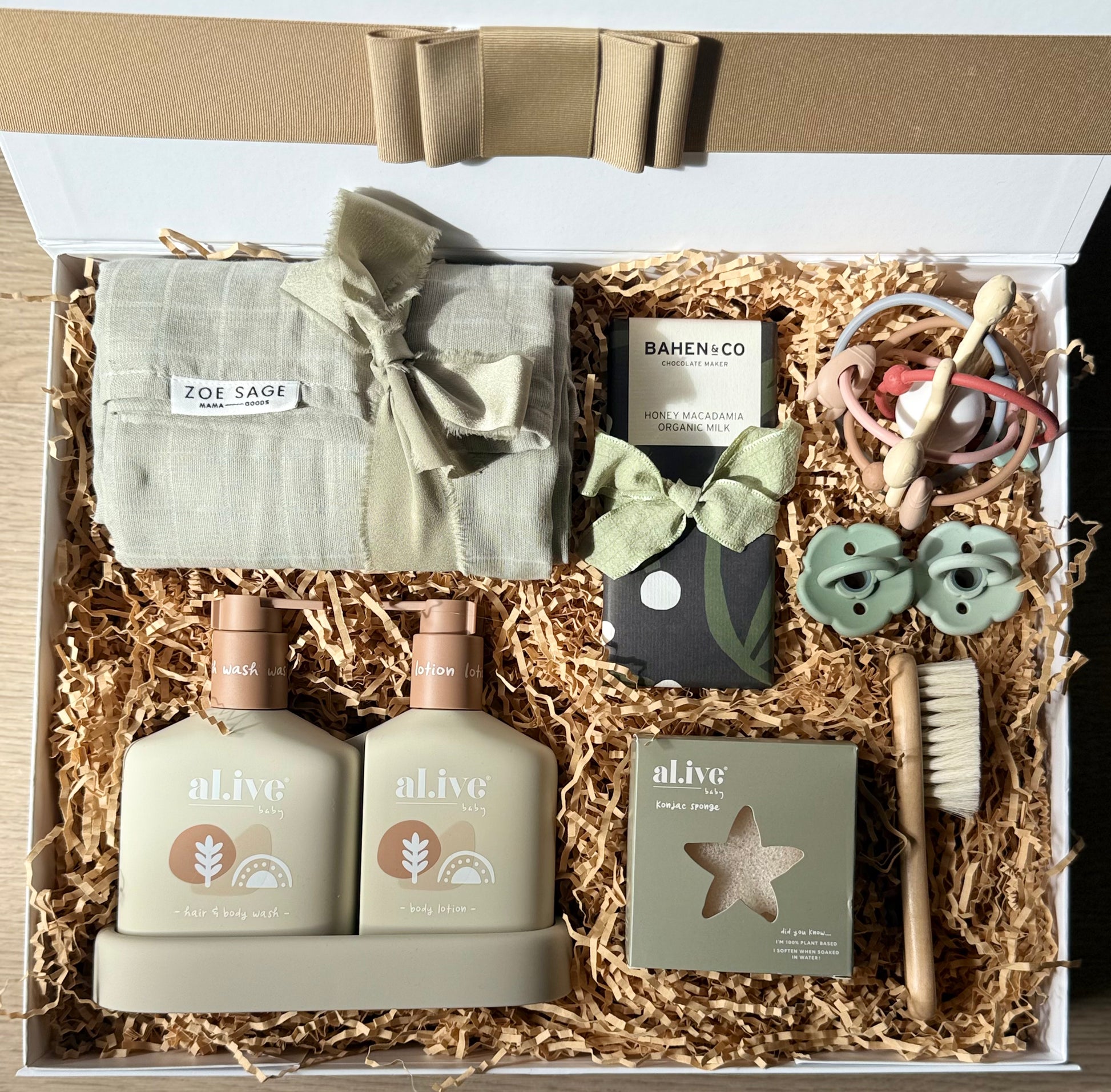The Baby Suite gift box featuring al.ive baby Hair & Body Duo, swaddle wrap, Bahen & Co chocolate, baby brush, konjac sponge, rattle teether toy, and Bebebiu dummies on shredded paper